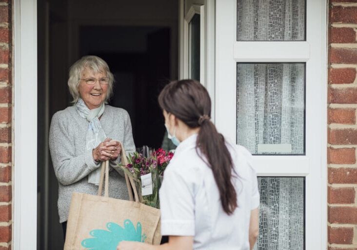 A female healthcare worker wearing a protective mask dropping off groceries and flowers at her grandmother's house as she stands at the door during the Coronavirus pandemic.